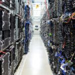 A brightly lit aisle in a data center, lined with racks of servers and neatly bundled cables on both sides, with blinking indicator lights and a concrete floor.