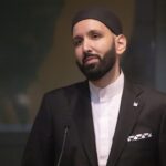 A man with a beard, wearing a black suit, white shirt, and black cap, stands in front of a microphone, appearing thoughtful. The background is dimly lit and out of focus.