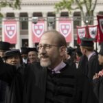 A man in academic regalia waves at a graduation ceremony, surrounded by graduates in caps and gowns with Harvard banners in the background.