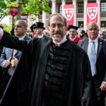 A man in academic robes waves at a graduation ceremony, surrounded by graduates and officials. Harvard banners and a crowd are visible in the background.
