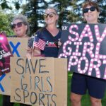 Three women stand outdoors holding signs reading SAVE GIRLS SPORTS and TWO XS NO EXCEPTIONS, showing support for girls sports. They smile and wear sunglasses, with trees and grass in the background.