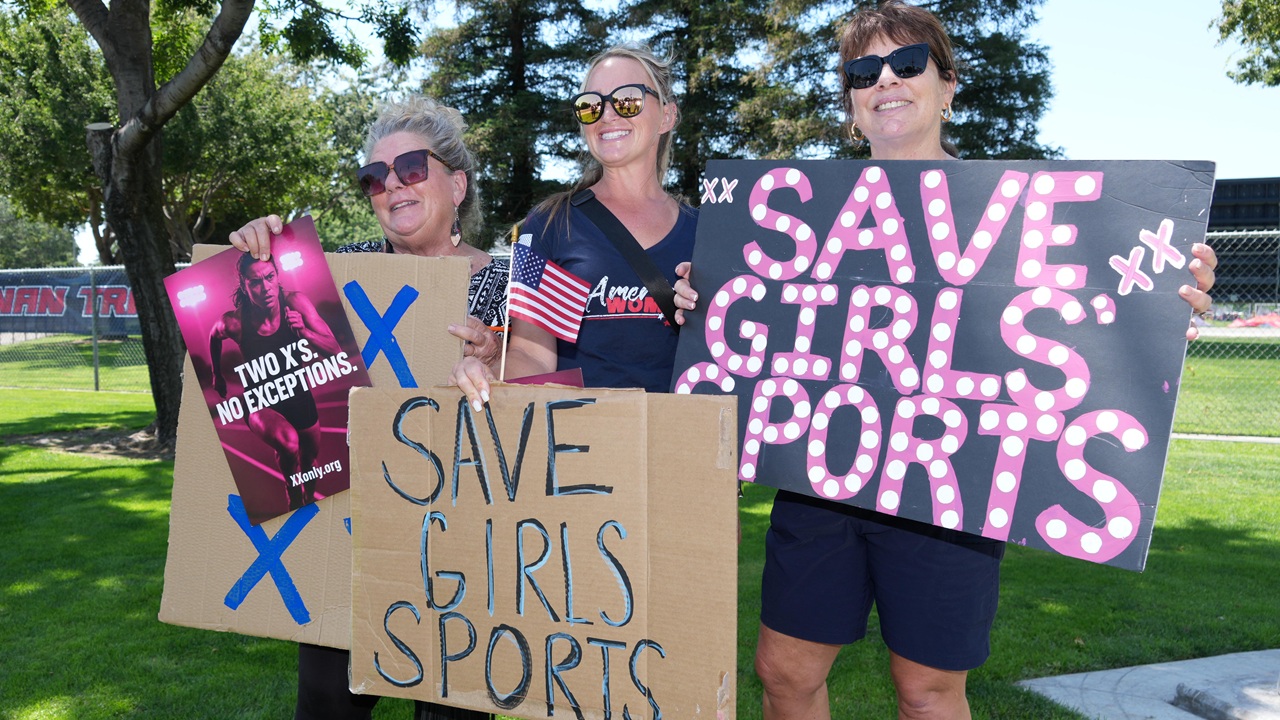 Home 4 Three women stand outdoors holding signs reading SAVE GIRLS SPORTS and TWO XS NO EXCEPTIONS, showing support for girls sports. They smile and wear sunglasses, with trees and grass in the background.