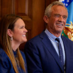 RFK Jr. in a blue suit and Gov. Sanders in a blue dress stand together indoors in front of an American flag.