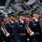 A group of uniformed soldiers marches in formation, carrying rifles with bayonets fixed, during a ceremonial parade.