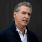 Governor Gavin Newsom, wearing a white shirt and dark jacket, speaks in front of a plain, dark background.