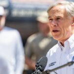 A man in a white shirt with a Texas emblem speaks at a podium outdoors, with people standing in the blurred background.