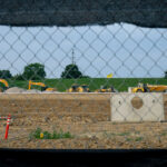 View of a construction site with excavators and trucks, seen through a chain-link fence and a black mesh barrier. Dirt, caution tape, and a concrete barrier are visible in the foreground.
