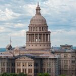 The Texas State Capitol building in Austin, with scaffolding on parts of its exterior, stands under a cloudy sky. The Texas flag is visible on the right side.