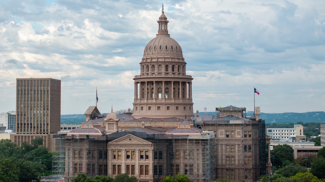 The Texas State Capitol building in Austin, with scaffolding on parts of its exterior, stands under a cloudy sky. The Texas flag is visible on the right side.