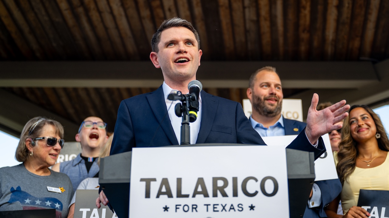 A man in a suit speaks at a podium with a microphone, holding a Talarico for Texas sign, surrounded by smiling supporters under a wooden roof.