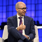 A man in a suit and tie, wearing glasses and a headset microphone, sits onstage with a blue geometric background, gesturing with his hand while speaking.