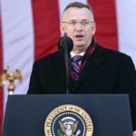 A man in a black coat and red-striped tie speaks at a podium with the U.S. presidential seal. Large American flag and gold eagle decorations are in the background.