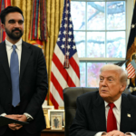 A man in a suit stands next to another man sitting at a desk in an office with U.S. flags, curtains, and framed photos in the background.