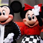 Mickey Mouse and Minnie Mouse, in themed outfits, stand side by side. Minnie waves while Mickey holds her hand. Both wear black, white, and red clothing with checkered details. A blurred person is behind them.