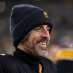 A smiling Aaron Rogers in a black beanie and black-and-gold jersey stands on the field, with blurred teammates and stadium lights in the background.