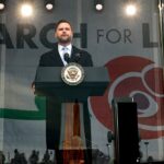 A man in a suit speaks at a podium with the Vice Presidents seal in front of a March for Life banner featuring a red rose graphic.