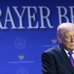 A man in a suit stands with his eyes closed in front of a blue backdrop with the words PRAYER BREAK partially visible.