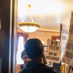 People holding protest signs against data centers indoors, with one sign featuring a large red slash through the words DATA CENTERS and others mentioning BIG TECH and demanding to keep every new data center out of Wisconsin.