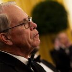 An older man wearing glasses and a tuxedo listens attentively at a formal event, with other people in tuxedos blurred in the background.