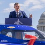 Transportation Secretary Sean Duffy speaking at a podium in front of a car