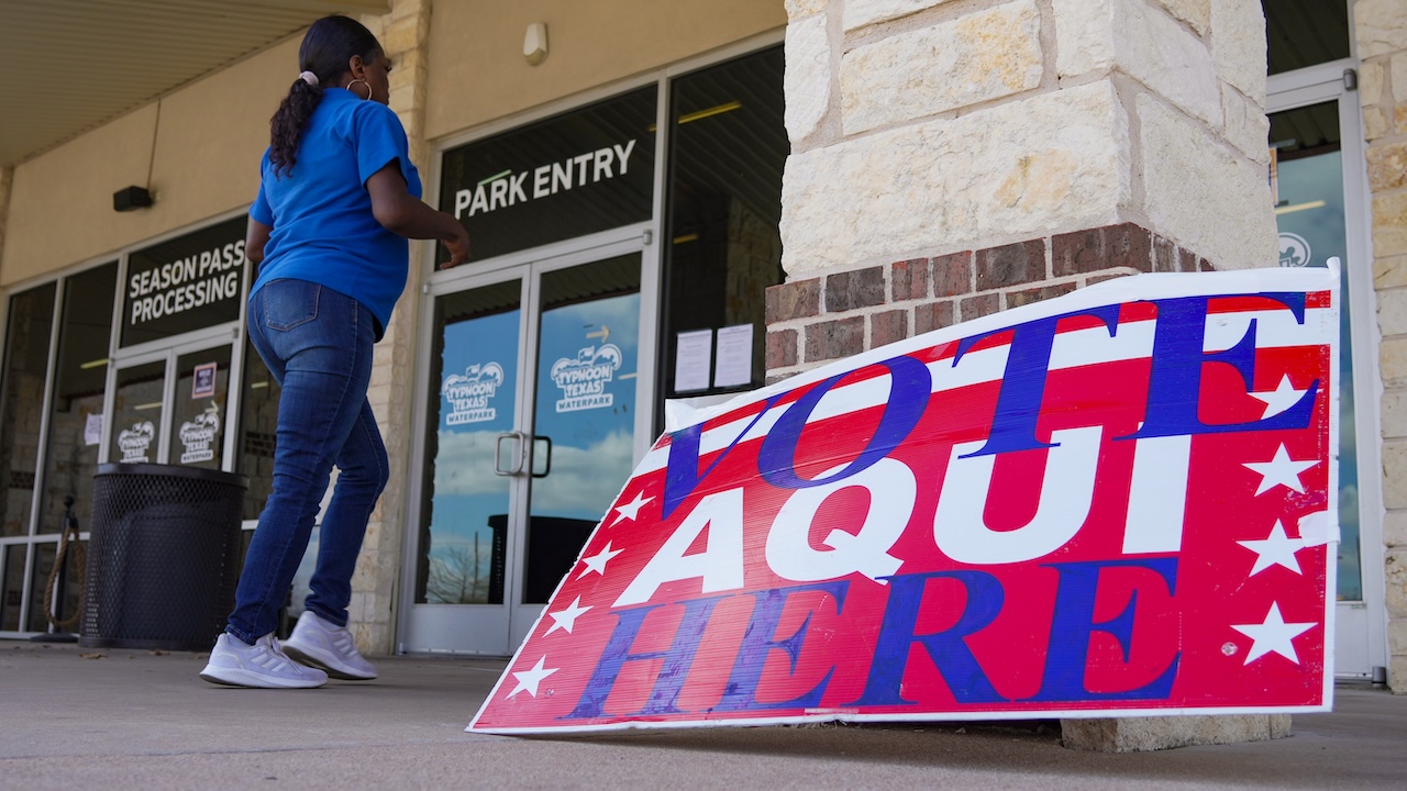 Home 2 A woman walks toward a building entrance with a bilingual VOTE AQUI HERE sign in red, white, and blue in the foreground.