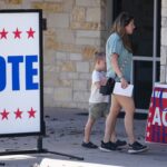 A woman and a child walk past large VOTE signs, one in English and one in Spanish, outside a stone building serving as a polling place.