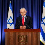 Benjamin Netanyahu stands at a podium with the Israeli emblem, flanked by two Israeli flags, delivering a speech about Israel in front of a blue curtain.
