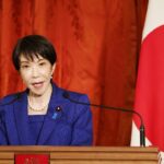 A woman in a blue suit speaks at a podium with microphones, standing beside a Japanese flag and a red curtain backdrop.