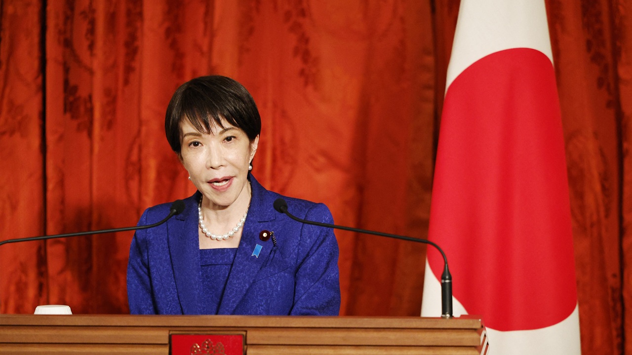 A woman in a blue suit speaks at a podium with microphones, standing beside a Japanese flag and a red curtain backdrop.