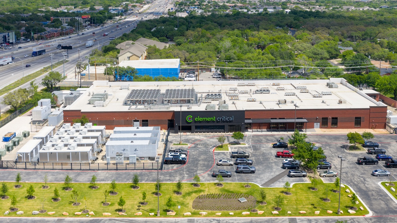 Aerial view of a large brick building labeled Element Critical with HVAC units on the roof, parking lot with cars, landscaped grass, and surrounding roads and trees.