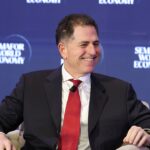 A man in a suit and red tie smiles while seated on stage at the Semafor World Economy event, with the events blue logo backdrop behind him.