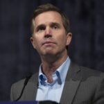 A man in a gray suit and blue shirt stands at a microphone, looking slightly upward. The background is dark and out of focus.