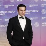 A man in a black tuxedo and bow tie stands in front of a purple and orange Breakthrough Prize backdrop, smiling with his hands in his pockets.