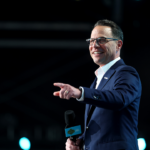 A man in a suit and glasses smiles while holding a microphone and pointing. The background is dark with blurred lights.