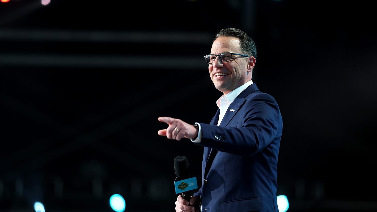 A man in a suit and glasses smiles while holding a microphone and pointing. The background is dark with blurred lights.