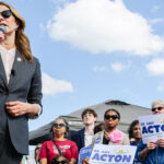 Dr. Amy Acton, wearing a dark suit, speaks into a microphone outdoors to a crowd holding Dr. Amy Acton for Governor signs.