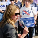 A woman in sunglasses speaks into a microphone at a rally. People behind her hold blue signs reading EDUCATORS FOR ACTION and DR. AMY ACTON FOR GOVERNOR. Some wear shirts supporting Amy Acton and Hambly.