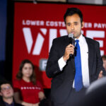 Republican Vivek Ramaswamy speaks in front of a crowd with a microphone in his hand and his red campaign backdrop behind him.