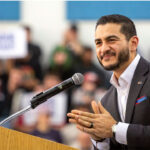 Abdul El-Sayed stands smiling at a podium in a dark suit.
