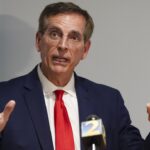 A man in a navy suit and red tie speaks with his hands raised in front of microphones at a press conference.