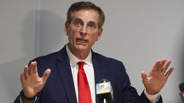 A man in a navy suit and red tie speaks with his hands raised in front of microphones at a press conference.