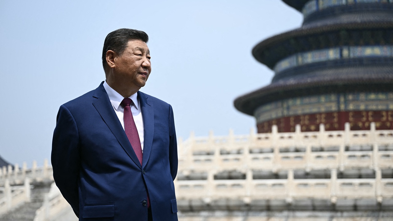 A man in a blue suit and red tie stands in front of an ornate, traditional Asian building with layered roofs and stone steps, under a clear sky.