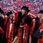 A group of graduates in caps and gowns celebrate outdoors, smiling and cheering. They wear sashes and sunglasses, with a stadium full of people in the background.