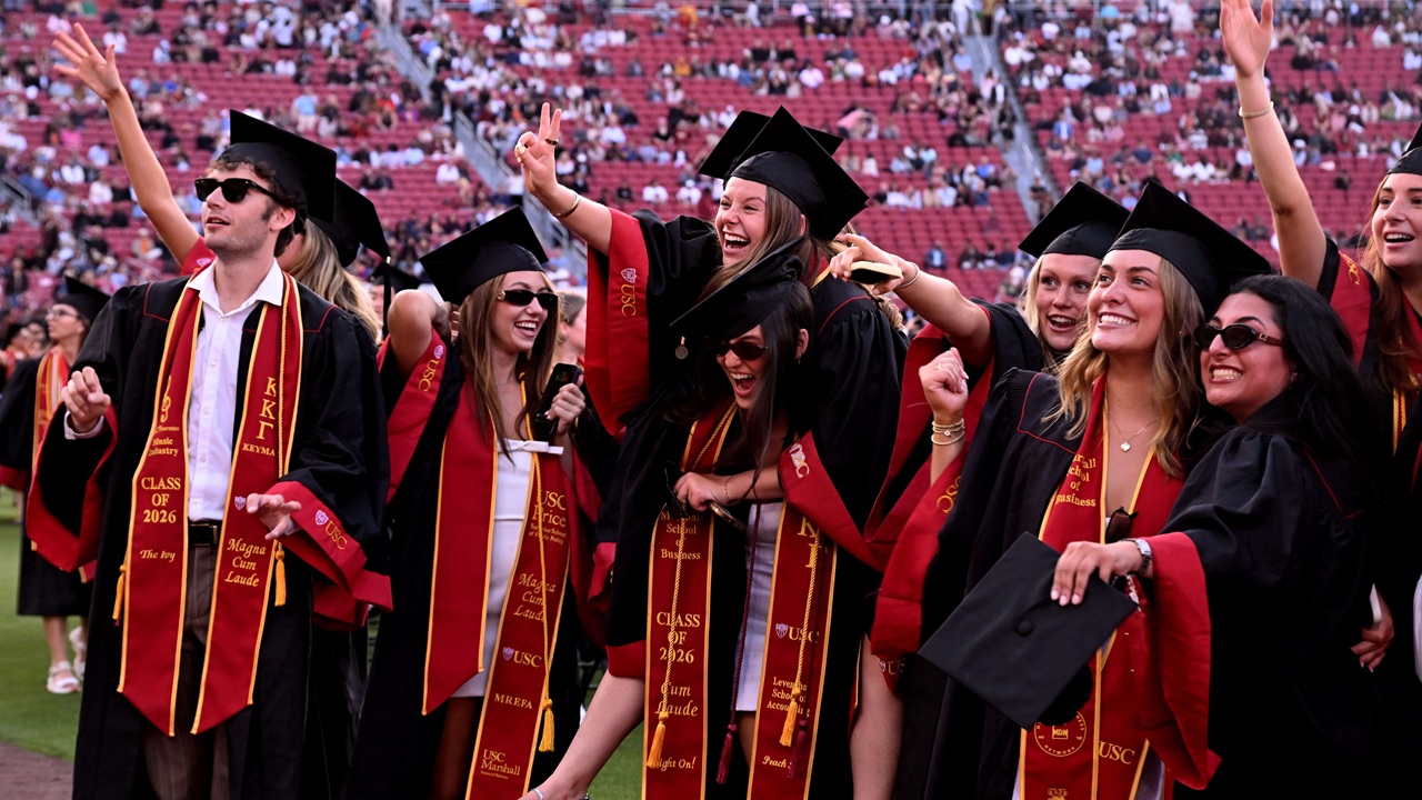 A group of graduates in caps and gowns celebrate outdoors, smiling and cheering. They wear sashes and sunglasses, with a stadium full of people in the background.