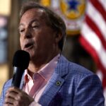 A man in a blue checkered blazer and pink shirt speaks into a microphone. An American flag and blurred background with signage are visible behind him.