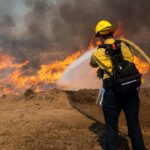 A firefighter points a fire hose towards brush that is caught on fire.