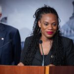 A woman smiles at a podium with microphones, wearing a black suit. An older man in a suit stands behind her. A blurred government building is in the background.