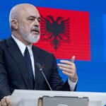 A man in a suit speaks at a podium with microphones. Behind him are the Albanian flag and the European Union flag on a blue background.