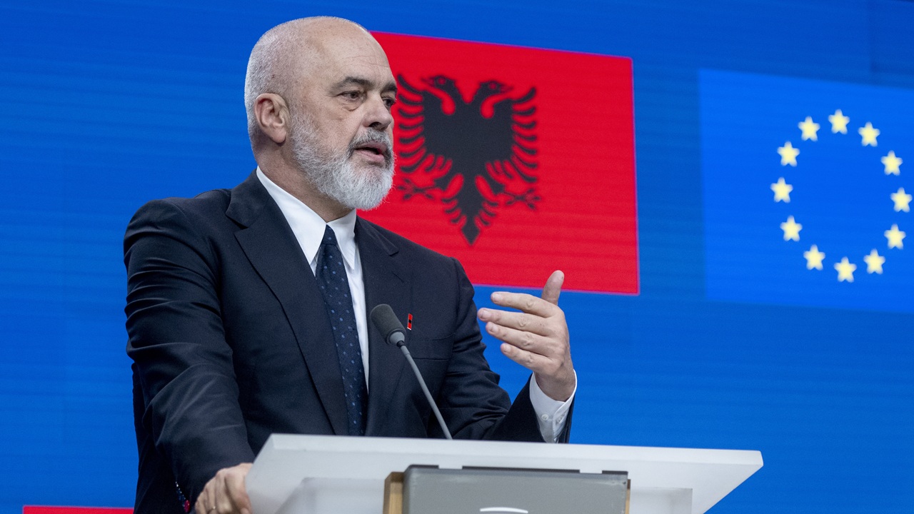 A man in a suit speaks at a podium with microphones. Behind him are the Albanian flag and the European Union flag on a blue background.