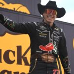A race car driver in a cowboy hat and sunglasses gives a thumbs up while standing in front of large Cracker Barrel 400 signs at a racetrack.